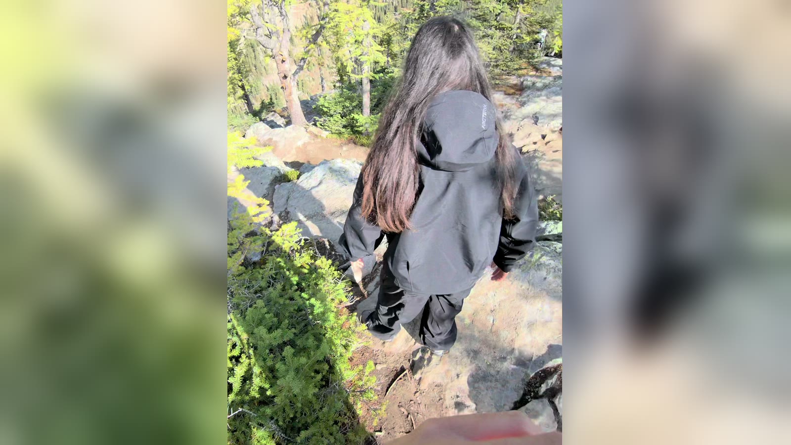 A person with long dark hair and a dark jacket, seen from behind, stepping down a rocky trail among pines; an upright clip over a blurred copy of itself